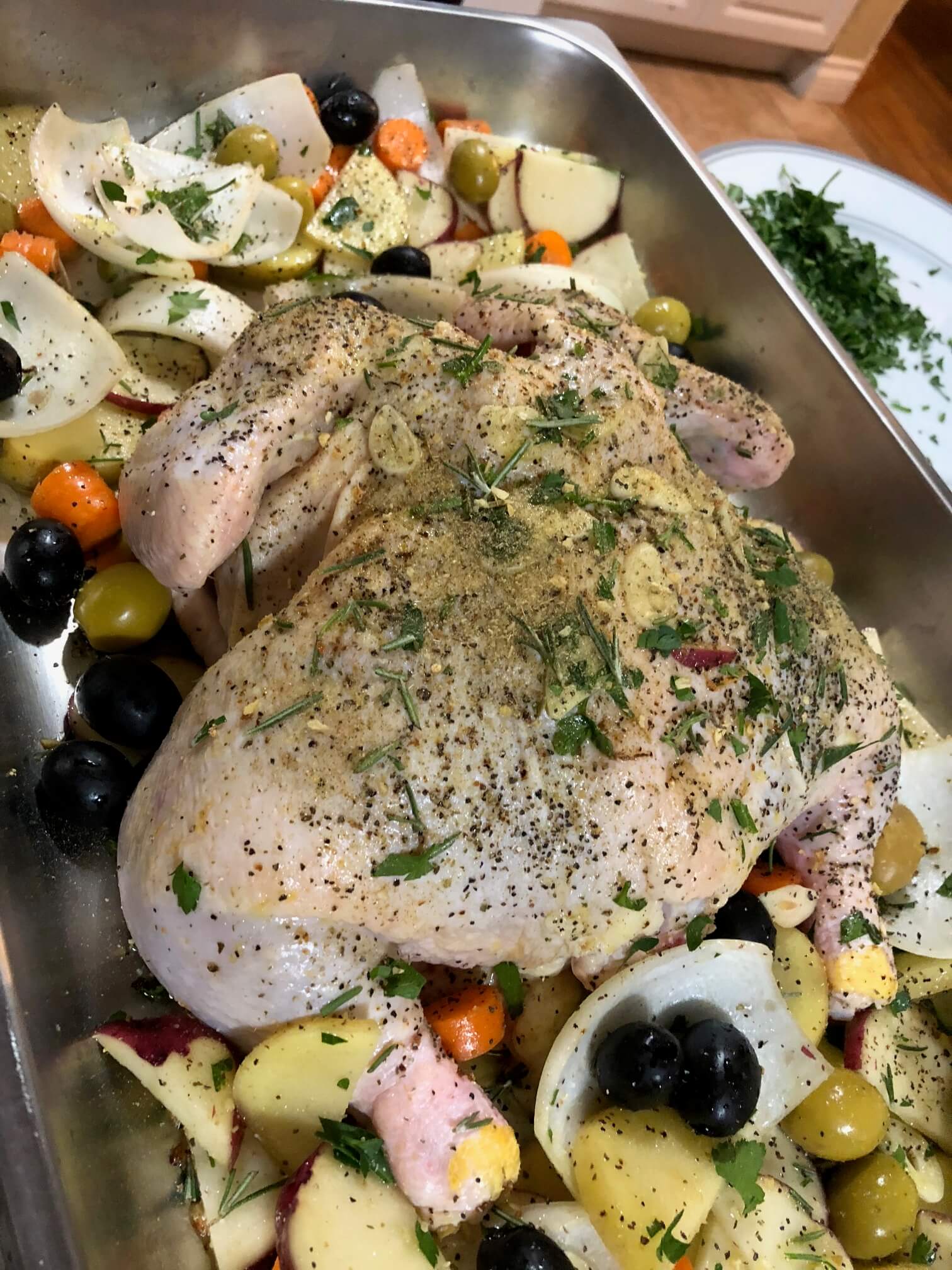 Close up of uncooked chicken with herbs and spices on a bed of vegetables in a silver pan
