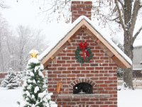 Red brick enclosed Casa 90 Pizza Oven with green wreath and red bow on front - Snow falling - small evergreen next to it