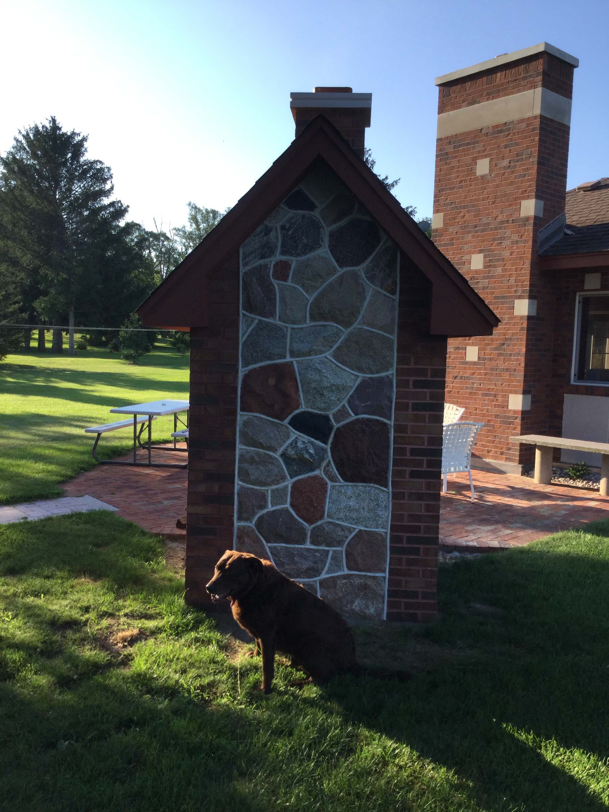 Dog standing in front of the backside of a brick and river rock pizza oven enclosure with a pointed roof