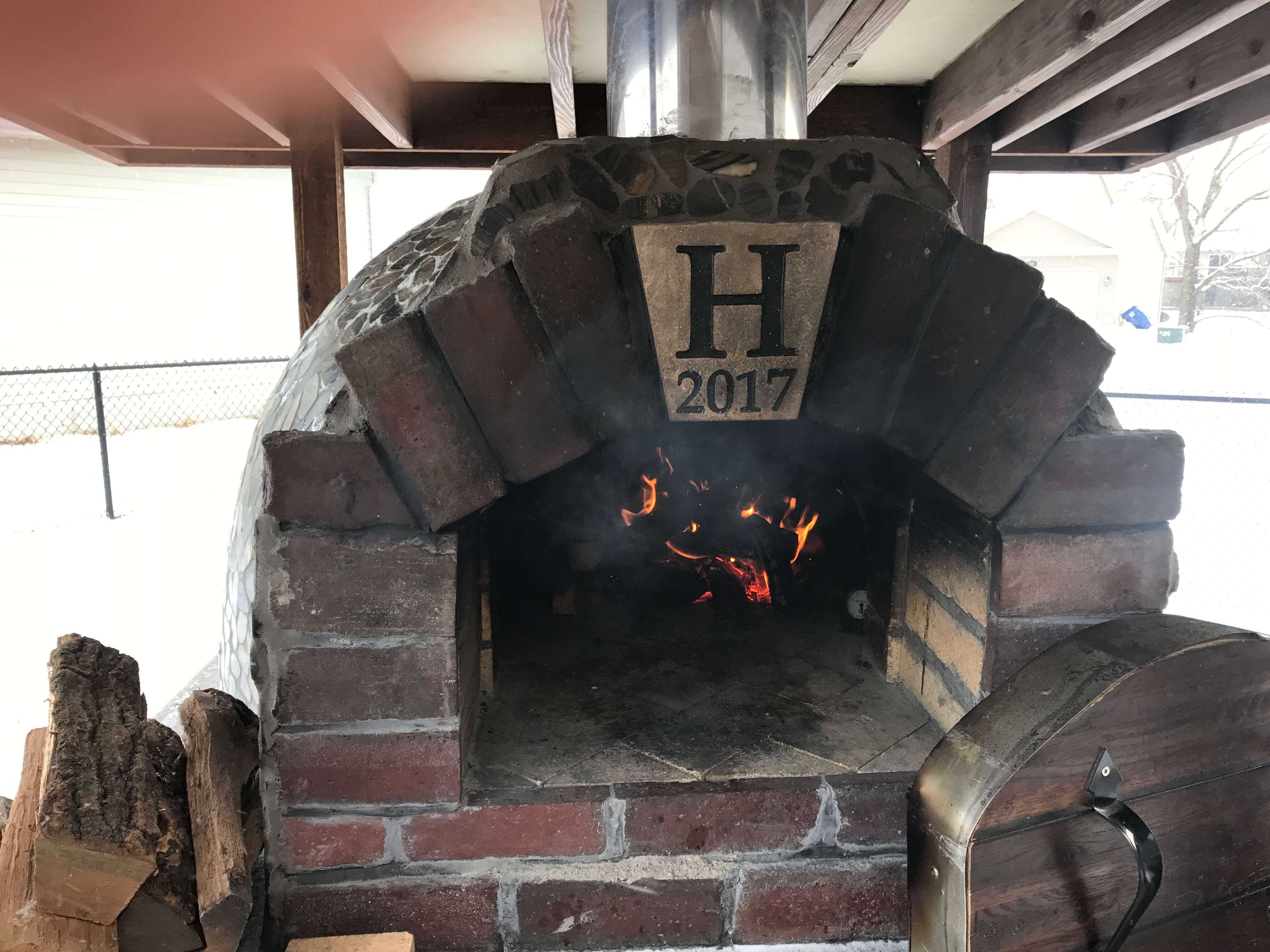 Dome shaped Pompeii brick pizza oven with fire burning - large H on the arch's capstone - snow in the background