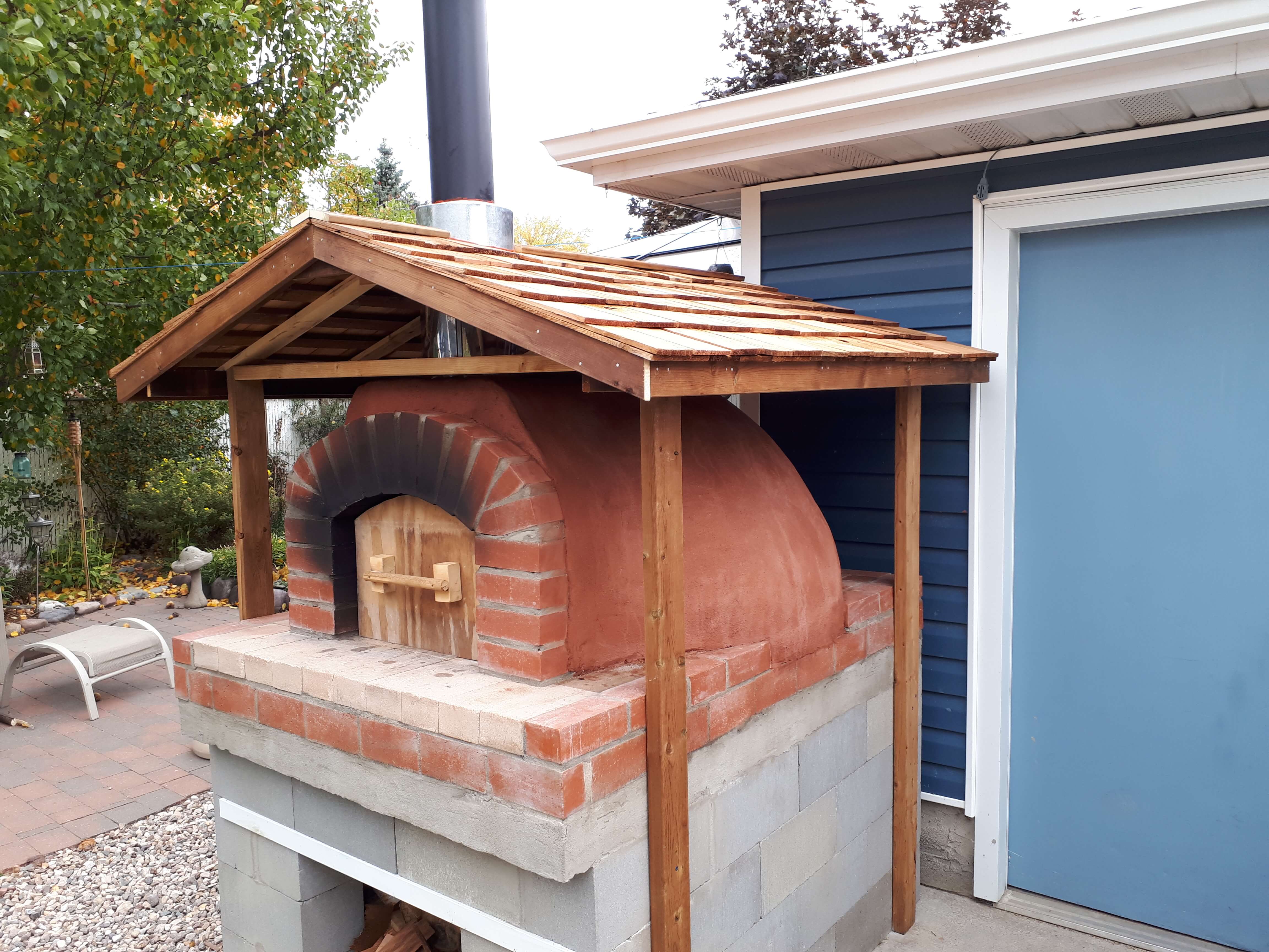 Red stucco pizza oven with brick arch and simple wooden door under a wooden roof with shingles on a cinderblock stand