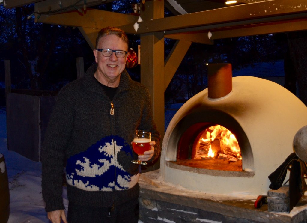 Smiling man in knit sweater holding a glass of beer in front of a burning cream-colored Primavera pizza oven