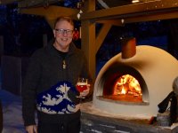 Smiling man in knit sweater holding a glass of beer in front of a burning cream-colored Primavera pizza oven