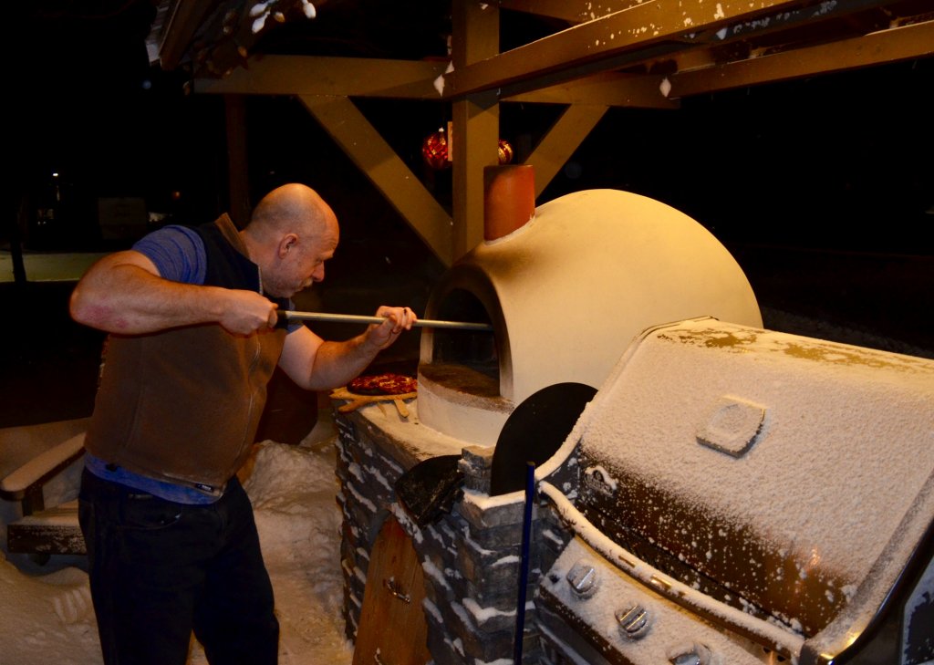 Side view of man with short sleeves and a pizza peel turning pizza in a Primavera oven on a covered patio with snow on the grill next to him