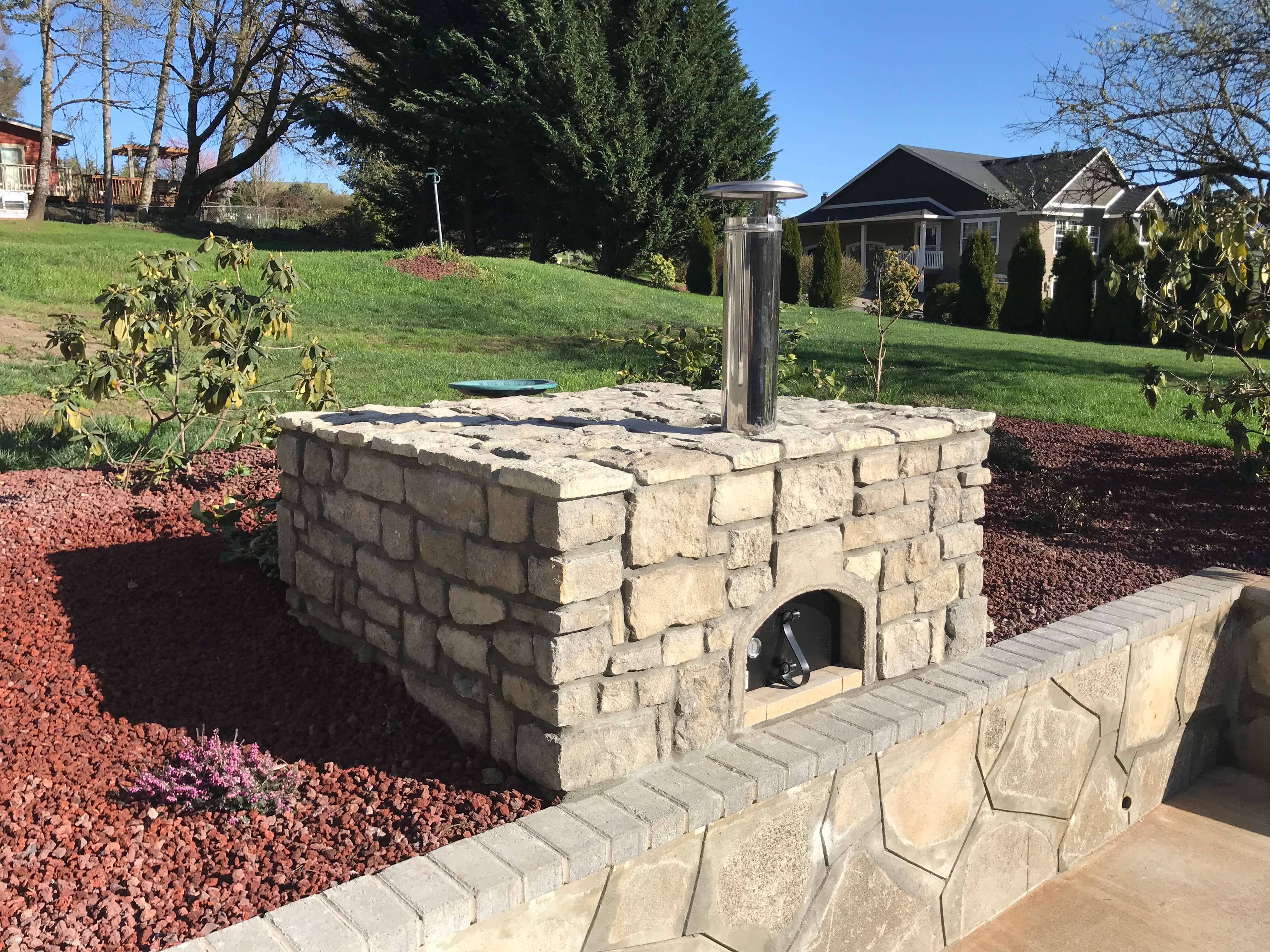 Side angle of stone pizza oven enclosure with black metal door and matching stone retaining wall