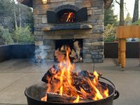 Black Metal Firepit in Foreground - Stone Enclosed Casa 110 Pizza Oven with Fire Burning and Metal Doors Open in the Background