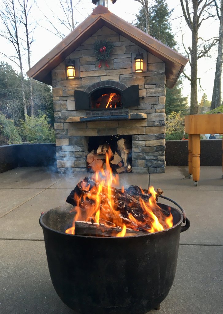 Black Metal Firepit in Foreground - Stone Enclosed Casa 110 Pizza Oven with Fire Burning and Metal Doors Open in the Background